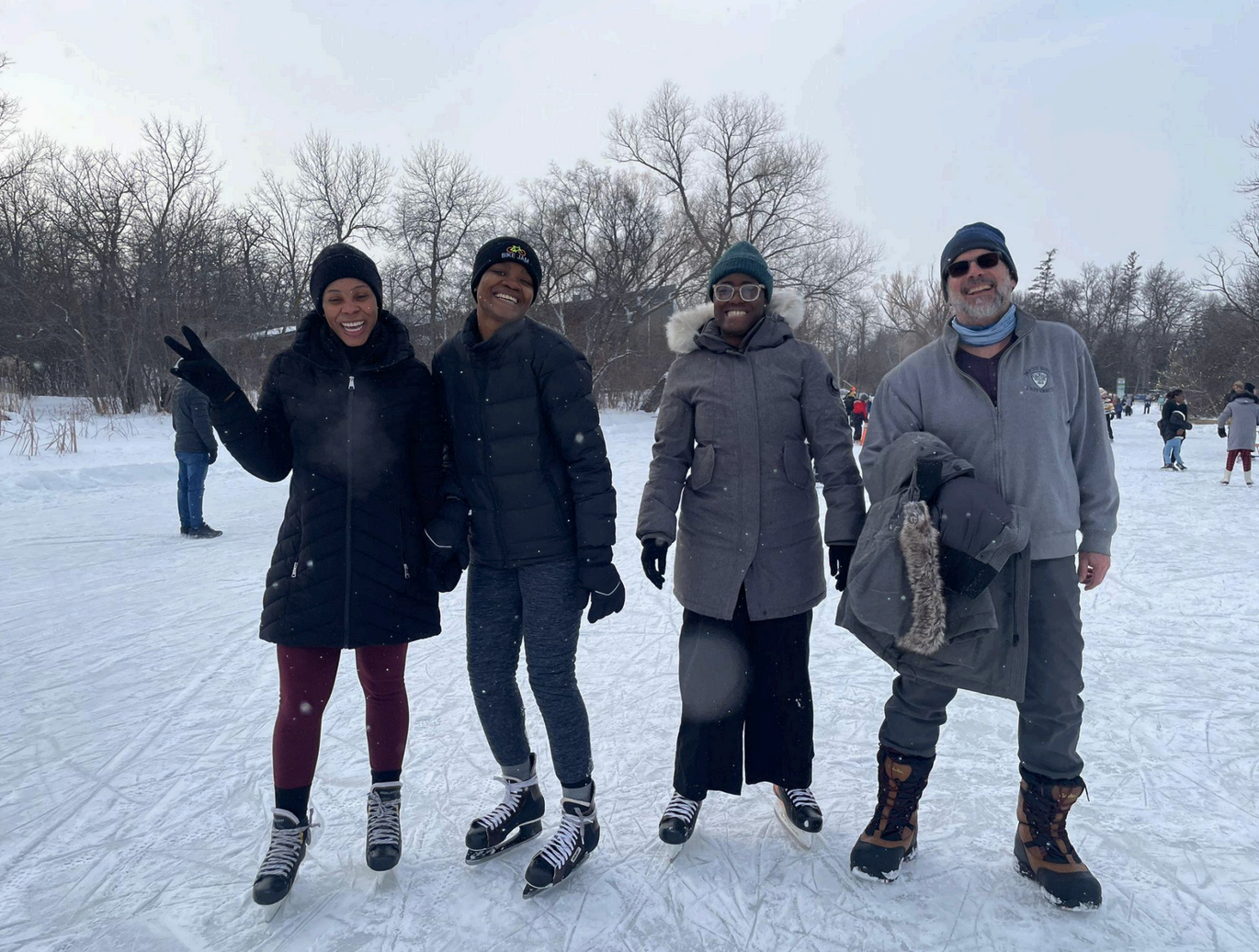 Three women wearing skates and one man wearing winter boots standing on the ice. All are smiling.